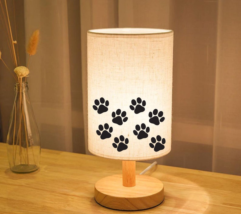 May include: A white linen lampshade with black paw prints, a wooden base, and a wooden stem. The lamp is on a wooden table.