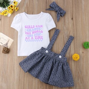 May include: A white t-shirt with the text "Girls Can Change The World One Step At A Time" in purple and blue lettering. The shirt is paired with a blue and white floral print skirt with suspenders and a pink bow headband.