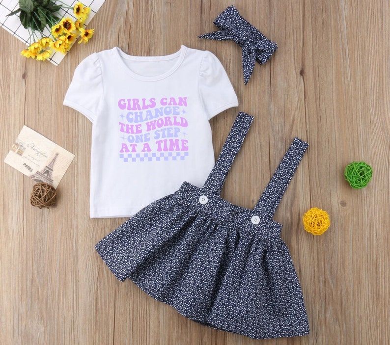 May include: A white t-shirt with the text "Girls Can Change The World One Step At A Time" in purple and blue lettering. The shirt is paired with a blue and white floral print skirt with suspenders and a pink bow headband.