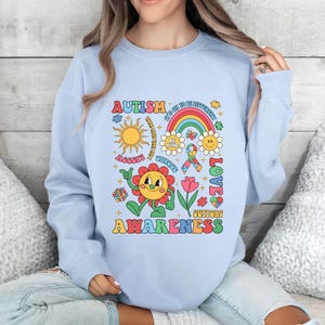 Pode incluir: Camisola azul claro com um design gr&aacute;fico colorido que apresenta uma flor, um arco-&iacute;ris, pe&ccedil;as de quebra-cabe&ccedil;a e o texto "Autism Awareness". O gr&aacute;fico tamb&eacute;m inclui o texto "It's OK to be Different", "Be Kind", "Unique", "Love" e "Support".