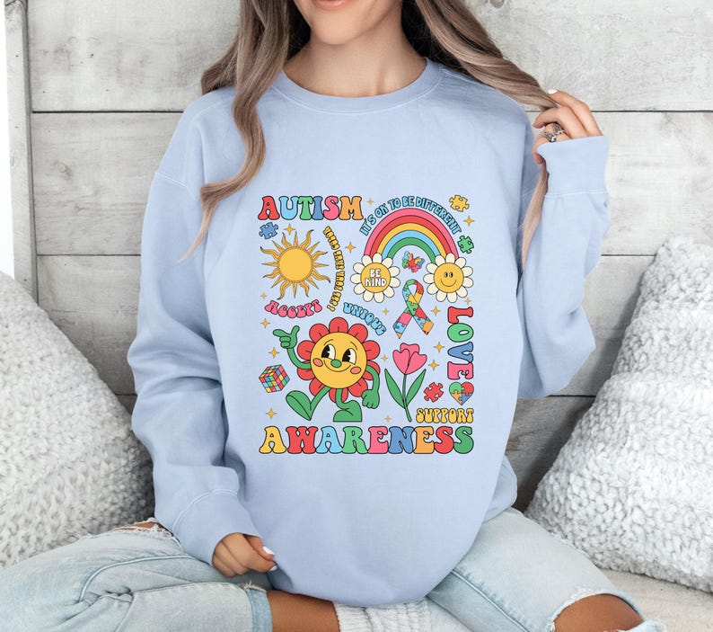 Pode incluir: Camisola azul claro com um design gr&aacute;fico colorido que apresenta uma flor, um arco-&iacute;ris, pe&ccedil;as de quebra-cabe&ccedil;a e o texto "Autism Awareness". O gr&aacute;fico tamb&eacute;m inclui o texto "It's OK to be Different", "Be Kind", "Unique", "Love" e "Support".