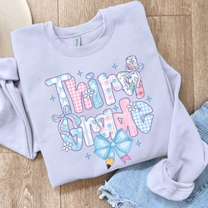 Puede incluir: Una sudadera lila claro con la frase "Third Grade" en un dise&ntilde;o colorido y estampado. Las letras est&aacute;n decoradas con cuadros, flores y lazos, y un gr&aacute;fico de l&aacute;piz. La sudadera est&aacute; doblada sobre una superficie de madera.