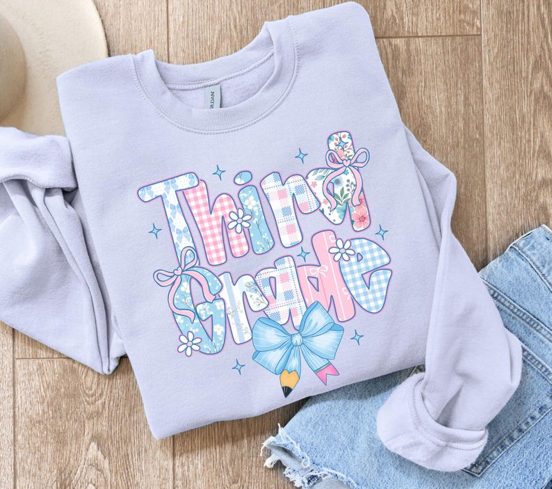 Puede incluir: Una sudadera lila claro con la frase "Third Grade" en un dise&ntilde;o colorido y estampado. Las letras est&aacute;n decoradas con cuadros, flores y lazos, y un gr&aacute;fico de l&aacute;piz. La sudadera est&aacute; doblada sobre una superficie de madera.