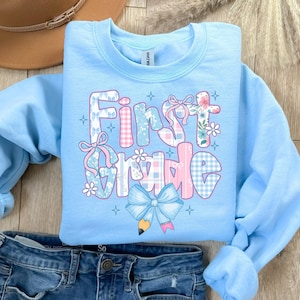 May include: Light blue sweatshirt with the words "First Grade" in a patchwork design. The letters are decorated with bows, flowers, and plaid patterns in pink, blue, and white. A large blue bow with a pencil graphic is at the bottom.