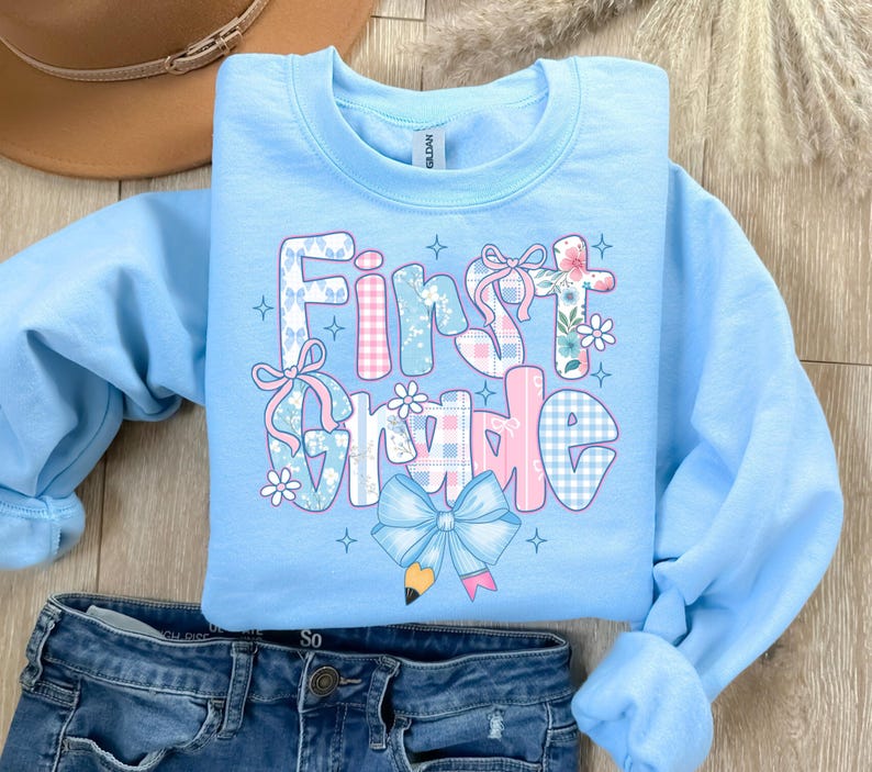 May include: Light blue sweatshirt with the words "First Grade" in a patchwork design. The letters are decorated with bows, flowers, and plaid patterns in pink, blue, and white. A large blue bow with a pencil graphic is at the bottom.