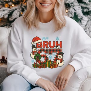 May include: White sweatshirt with a Christmas-themed graphic. The graphic includes the word "BRUH" in red letters with a Santa hat, the number "67" in a red and black plaid pattern, snowmen, snowflakes, and wrapped gifts.