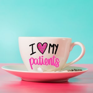 Puede incluir: Una taza de caf&eacute; blanca con un coraz&oacute;n rosa y el texto "Amo a mis pacientes" en letras negras.