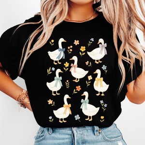 May include: Black t-shirt featuring a pattern of white geese with colorful bows around their necks. The geese are surrounded by small flowers and leaves in various colors. A charming and whimsical design.