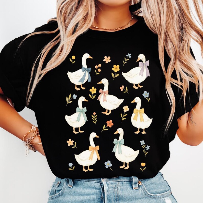 May include: Black t-shirt featuring a pattern of white geese with colorful bows around their necks. The geese are surrounded by small flowers and leaves in various colors. A charming and whimsical design.