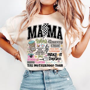 May include: A white t-shirt with a black graphic that says "Mama" and features a variety of colorful illustrations, including a coffee cup, a camera, a donut, and a lightning bolt. The text on the graphic says "Work Grocery Store, School Vacations Appointments Doctor Visits, Field Trips Playground, Make It Happen, Kitchen Practice Sport Games, The Motherhood Tour."