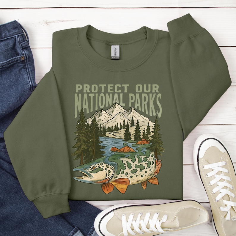 May include: Olive green sweatshirt with the text "PROTECT OUR NATIONAL PARKS" above a graphic of a fish, river, and mountains. The fish has a spotted pattern with orange fins. The sweatshirt is paired with blue jeans and white sneakers.