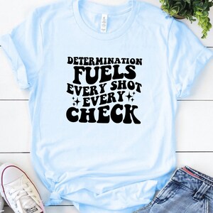 May include: Light blue t-shirt with black text that reads "Determination fuels every shot every check".