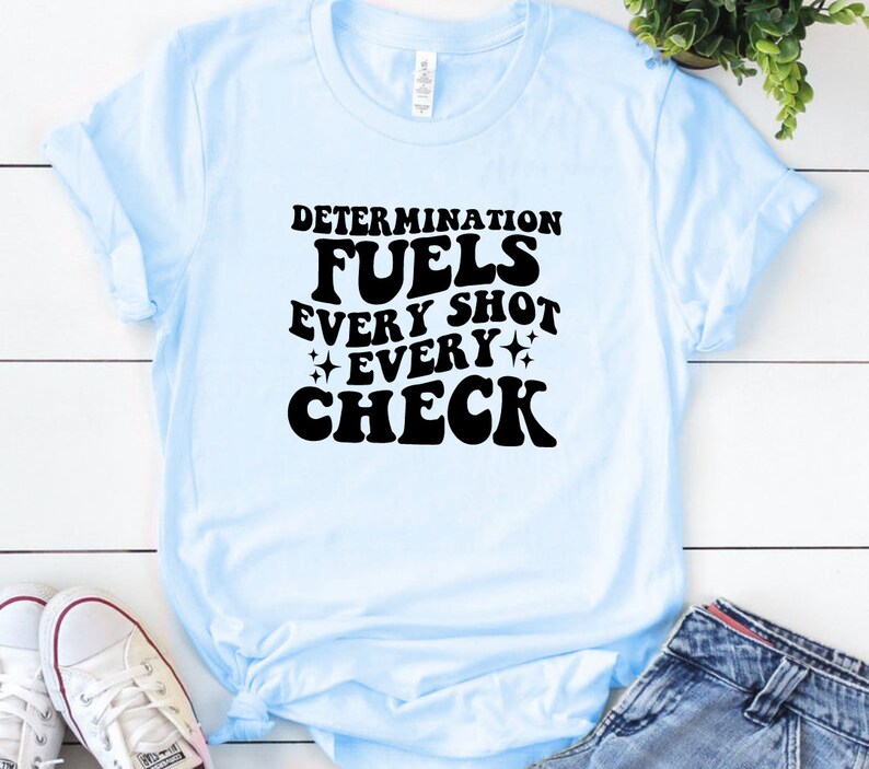 May include: Light blue t-shirt with black text that reads "Determination fuels every shot every check".