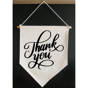 May include: A white canvas banner with a black cursive inscription that reads "Thank you". The banner is hanging from a string and has a wooden dowel rod at the top.