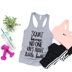 K&ouml;nnte beinhalten: Ein graues Tanktop mit dem Text "SQUAT because NO ONE RAPS ABOUT little butts" wird mit einem rosa Sport-BH, schwarzen Leggings, einer Wasserflasche, einer kleinen Pflanze und zwei lila Kurzhanteln gezeigt. Die Artikel sind auf einer wei&szlig;en Oberfl&auml;che angeordnet.
