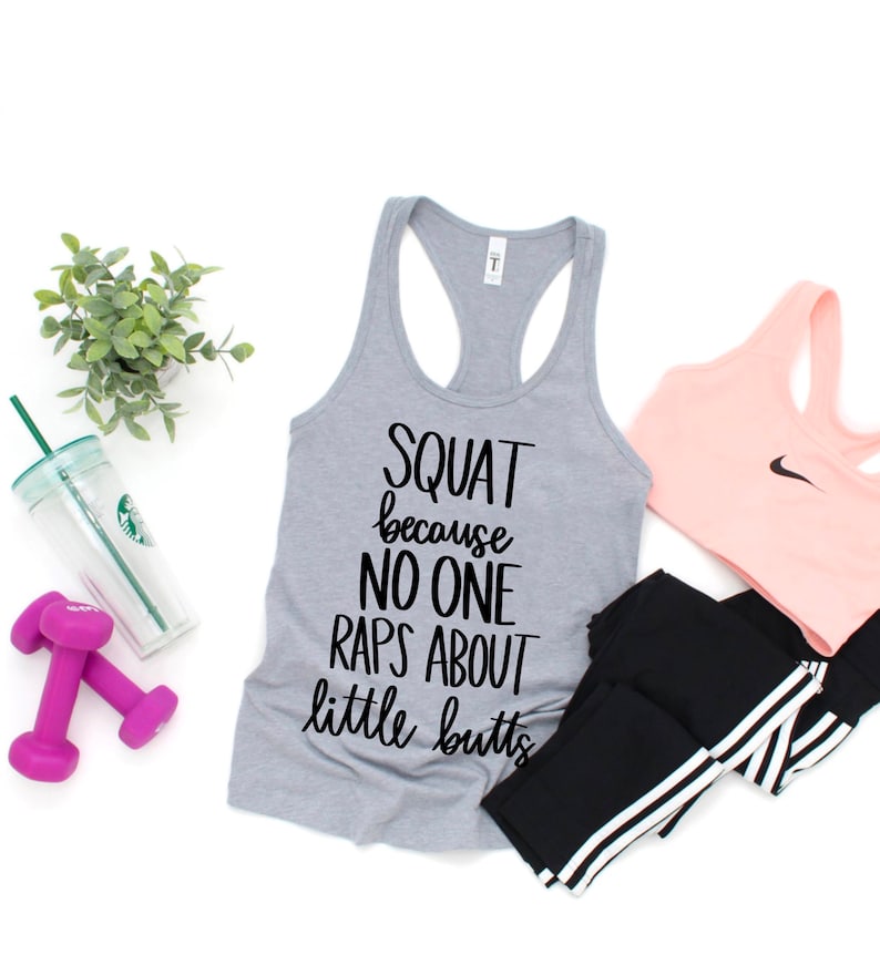 K&ouml;nnte beinhalten: Ein graues Tanktop mit dem Text "SQUAT because NO ONE RAPS ABOUT little butts" wird mit einem rosa Sport-BH, schwarzen Leggings, einer Wasserflasche, einer kleinen Pflanze und zwei lila Kurzhanteln gezeigt. Die Artikel sind auf einer wei&szlig;en Oberfl&auml;che angeordnet.