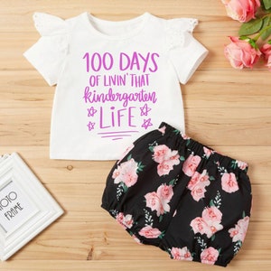May include: White short-sleeved shirt with a pink graphic that says "100 Days of Livin' That Kindergarten Life" with stars around the word "Life". The shirt has a white lace trim on the sleeves. Black and white floral print shorts.