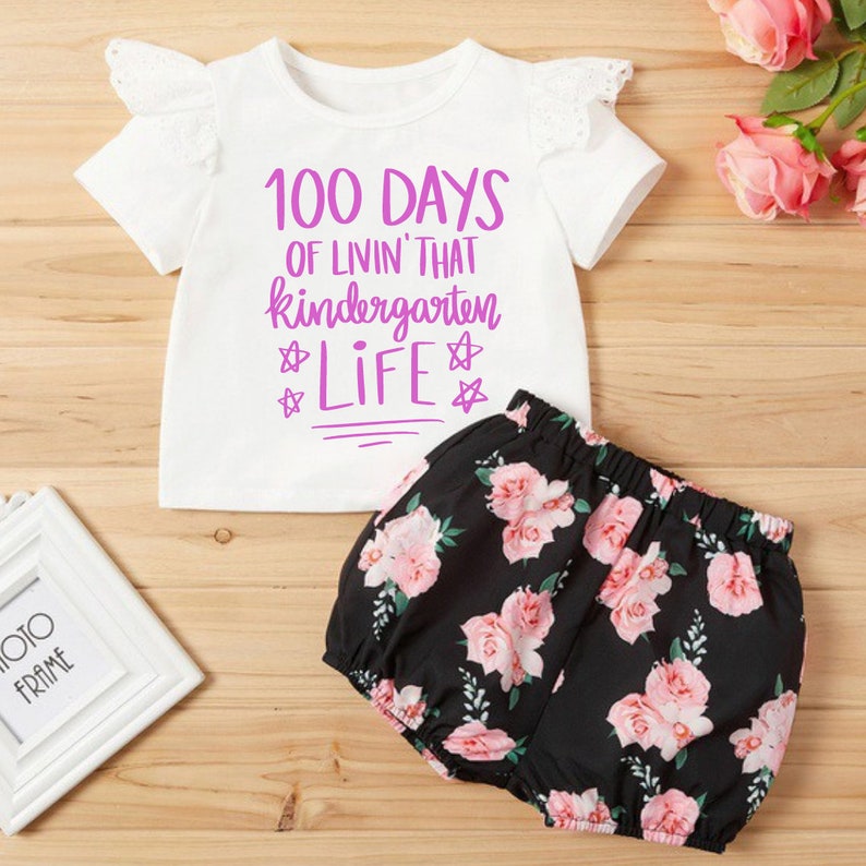 May include: White short-sleeved shirt with a pink graphic that says "100 Days of Livin' That Kindergarten Life" with stars around the word "Life". The shirt has a white lace trim on the sleeves. Black and white floral print shorts.