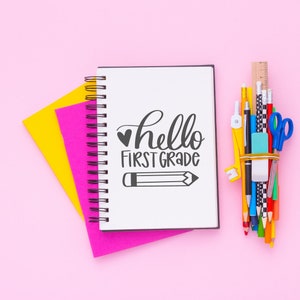 May include: A pink and yellow notebook with a spiral binding. The notebook has the words "hello first grade" written on the front. A bunch of colorful pencils, pens, and a ruler are on the right side of the image.