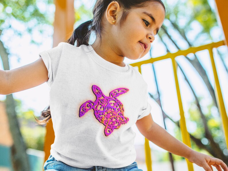 K&ouml;nnte beinhalten: Ein wei&szlig;es T-Shirt mit einem pinkfarbenen und violetten Muster einer Meeresschildkr&ouml;te. Die Schildkr&ouml;te ist nach rechts im Bild ausgerichtet.