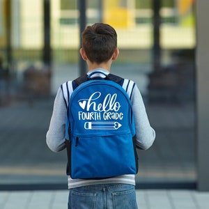 以下が含まれることがあります： 白いハートと「hello fourth grade」の文字、白い鉛筆のグラフィックが描かれた青いバックパック