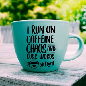 May include: A light blue ceramic coffee mug with a black graphic that reads "I run on caffeine chaos and cuss words *!@#".