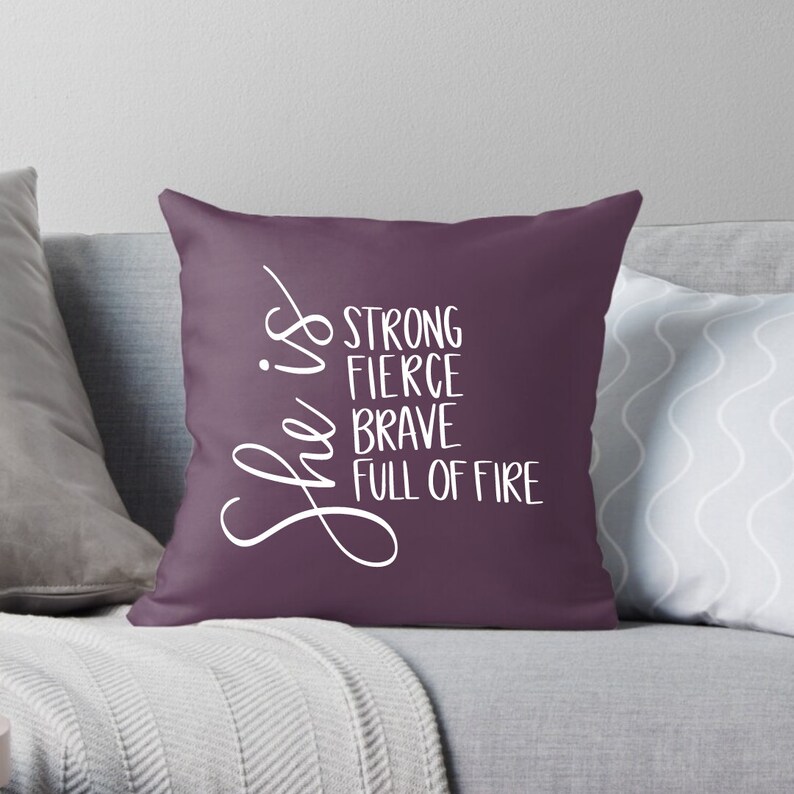 May include: Purple throw pillow with white text that reads "She is strong, fierce, brave, full of fire".