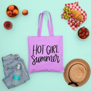 May include: Purple tote bag with the text "HOT GIRL Summer" in black lettering. The bag is surrounded by various summer-themed items, including fruit, a straw hat, and a picnic blanket.