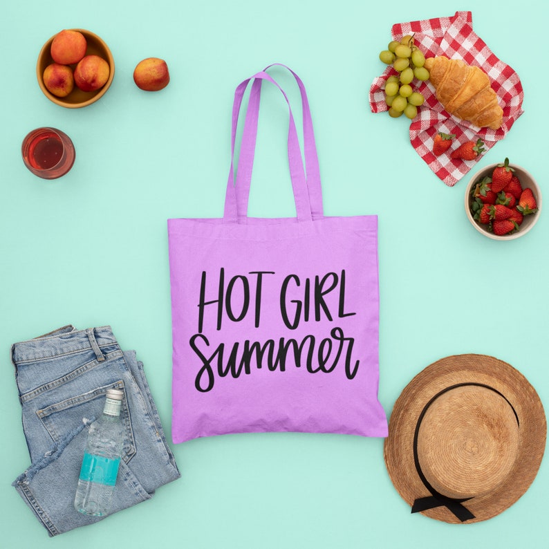 May include: Purple tote bag with the text "HOT GIRL Summer" in black lettering. The bag is surrounded by various summer-themed items, including fruit, a straw hat, and a picnic blanket.