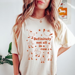 May include: Cream-colored t-shirt featuring a whimsical design of ducks and stars in a warm brown hue. The text "Definitely not all in a row" is centered below the duck illustrations, creating a playful and lighthearted aesthetic.