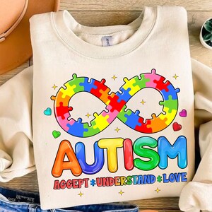 Pode incluir: Uma camisa de moletom cor creme com um s&iacute;mbolo de infinito arco-&iacute;ris feito de pe&ccedil;as de quebra-cabe&ccedil;a e a palavra "AUTISMO" em letras arco-&iacute;ris. O texto abaixo diz "ACCEPT * UNDERSTAND * LOVE".
