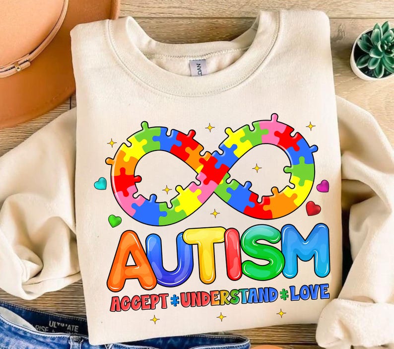 Pode incluir: Uma camisa de moletom cor creme com um s&iacute;mbolo de infinito arco-&iacute;ris feito de pe&ccedil;as de quebra-cabe&ccedil;a e a palavra "AUTISMO" em letras arco-&iacute;ris. O texto abaixo diz "ACCEPT * UNDERSTAND * LOVE".