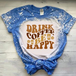 May include: A white t-shirt with a blue bleach splattered design. The shirt has a brown graphic that says "Drink Coffee Be Happy" with coffee cup illustrations.