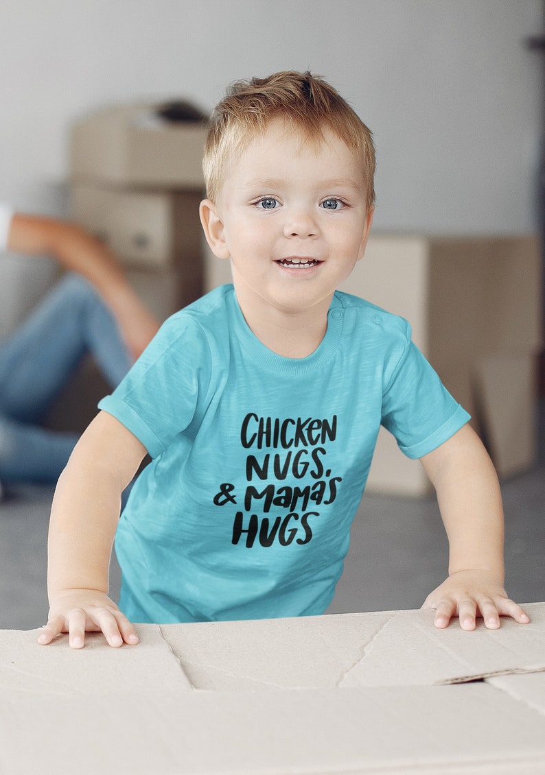 May include: A young child wearing a turquoise blue t-shirt with the text "Chicken Nugs. & Mamas Hugs" printed in black.