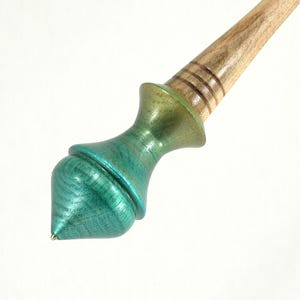 May include: A wooden tool with a tapered handle featuring three horizontal grooves and a bulbous, acorn-shaped end finished in a metallic teal and green gradient. The wood grain is visible throughout the natural tan handle section.