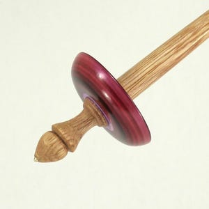 May include: A drop spindle featuring a long, light-colored wooden shaft and a smooth, deep burgundy whorl. The bottom of the spindle terminates in a carved, acorn-shaped wooden tip with a small, pointed metal end at the very base.