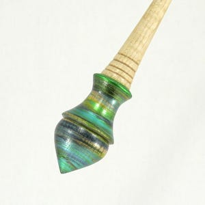 Puede incluir: Un palillo de pelo de madera con una cabeza decorativa iridiscente. La cabeza presenta una mezcla de tonos verdes, azules y dorados, con un brillo metálico. La parte superior del palillo es de color madera natural, y la cabeza tiene forma redondeada y cónica.