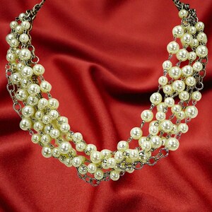 May include: A multi-strand necklace featuring faux pearls and silver-tone chains. The necklace is composed of several strands of off-white, round pearls, connected by delicate chains. The jewelry is displayed on a red satin background.