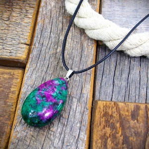Puede incluir: Un colgante en forma de lágrima con una vibrante piedra preciosa verde y púrpura, suspendido de un collar de cordón negro. La piedra preciosa está unida a un cierre de metal plateado. El fondo es una superficie de madera rústica.