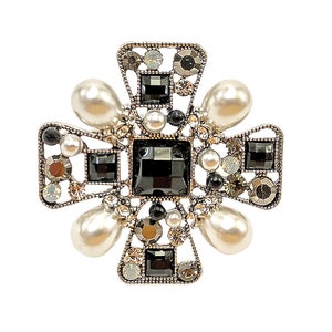 Op de afbeelding: Een zilveren broche in de vorm van een kruis met zwarte en witte strass steentjes en nepparels.