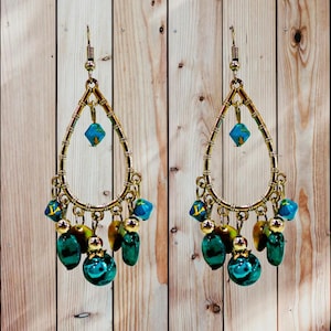 May include: A pair of gold-toned teardrop earrings with turquoise and green beads. The earrings have a decorative design with multiple dangling beads and a hook closure. The background is a light brown wooden surface.