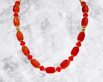 May include: A necklace featuring a mix of red and orange beads. The necklace has a gold-colored chain and small gold-colored beads interspersed between the larger beads. The beads are oval and round, creating a unique design.