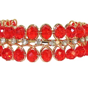 Puede incluir: Un brazalete de alambre dorado con cuentas de vidrio rojas y diamantes de imitación transparentes.
