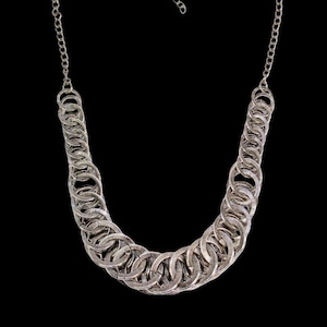 May include: A silver-toned necklace featuring a series of interlocking circular links that form a wide, curved collar shape. The chain is composed of multiple textured rings connected together, creating a dense, layered appearance with a metallic finish.