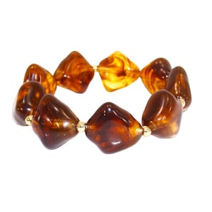 Puede incluir: Una pulsera elástica con cuentas de resina pulida de color ámbar irregular, con tonos marrones oscuros y dorados. Las cuentas están separadas por pequeñas cuentas espaciadoras redondas de metal en tono dorado brillante.