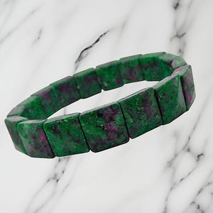 May include: A stretch bracelet composed of rectangular, polished beads made of green zoisite with embedded patches of deep magenta ruby. The beads are arranged in a continuous circular loop, showcasing a natural mottled texture and color variation.