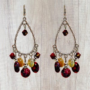 May include: Silver-toned teardrop earrings with dangling red, yellow, and purple beads. The earrings have a textured frame and feature a variety of bead shapes, including faceted squares and round orbs. The earrings are designed for pierced ears.