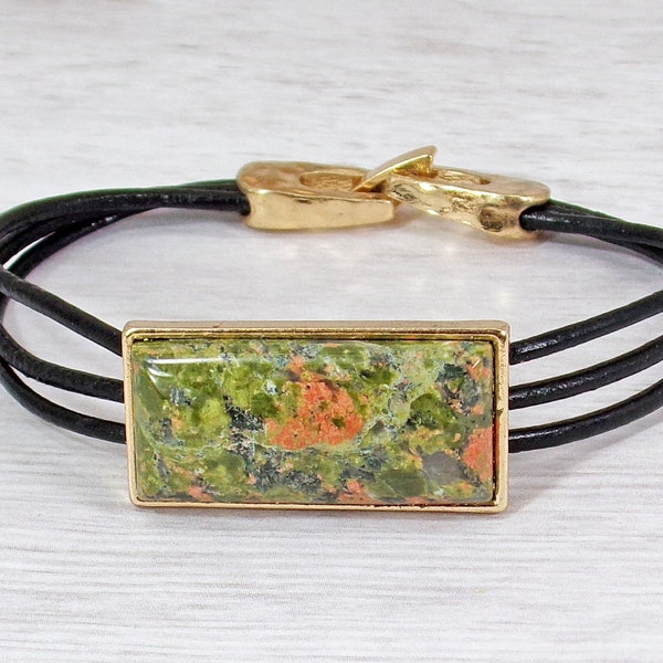 Green and pink unakite agate stone bracelet with a rectangle cut stone in a gold setting attached to three rows of black leather cord.