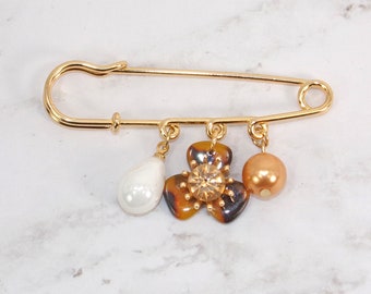Safety pin brooch with charm beads designed with a gold safety pin, a white pearl bead, tortoise color flower charm, and beige pearl