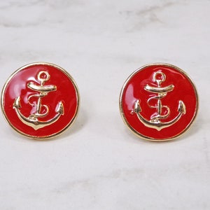 May include: A pair of red and gold anchor earrings. The anchors are gold and the background is red.